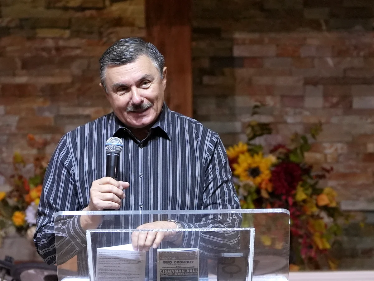 Mike Cousineau smiling as he speaks from the clear pulpit at New Hope, with autumn flowers behind him.