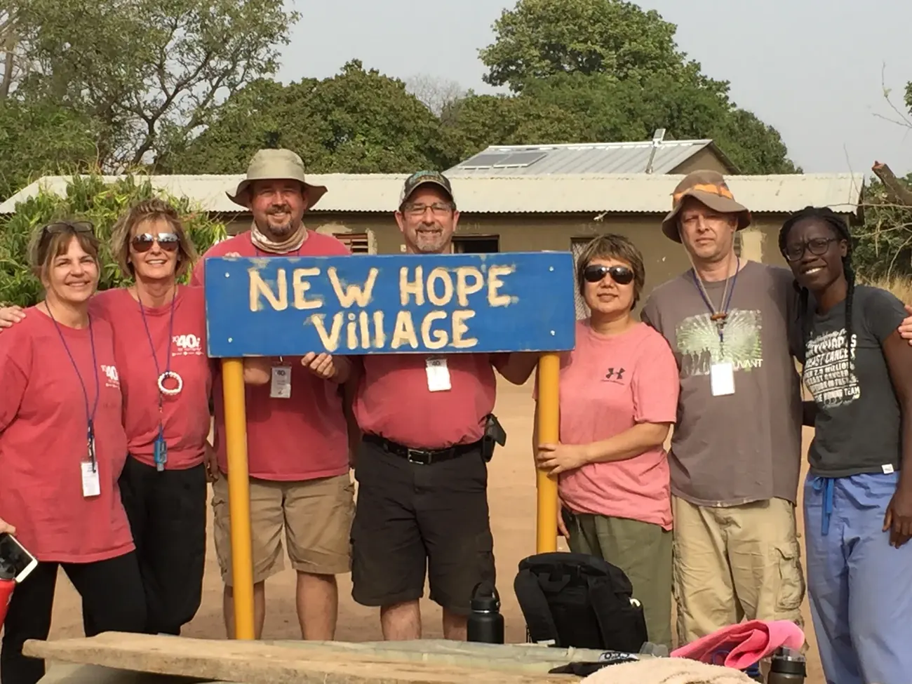 Seven people stand together behind a hand-painted blue sign that reads New Hope Village.