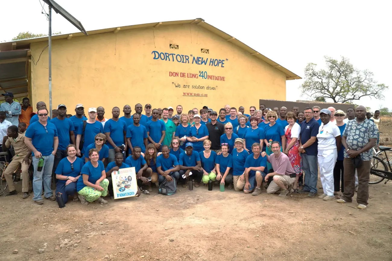 A large group in matching blue shirts stands in front of a yellow building with a painted sign that reads Dortoir New Hope.