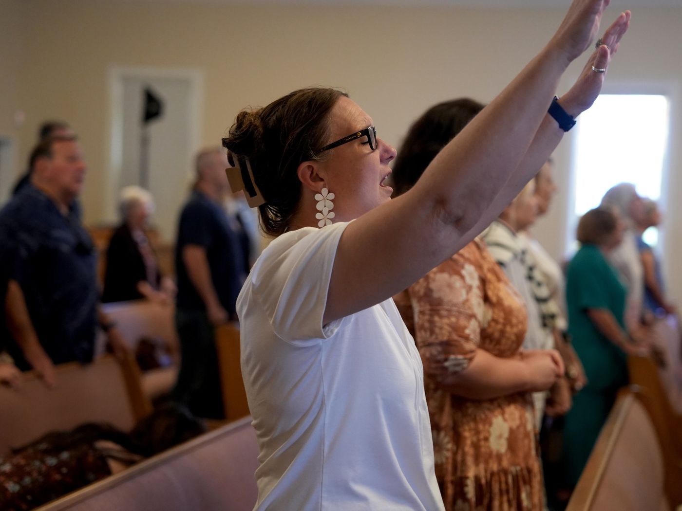 A woman singing with one hand raised during worship, others standing in the pews around her