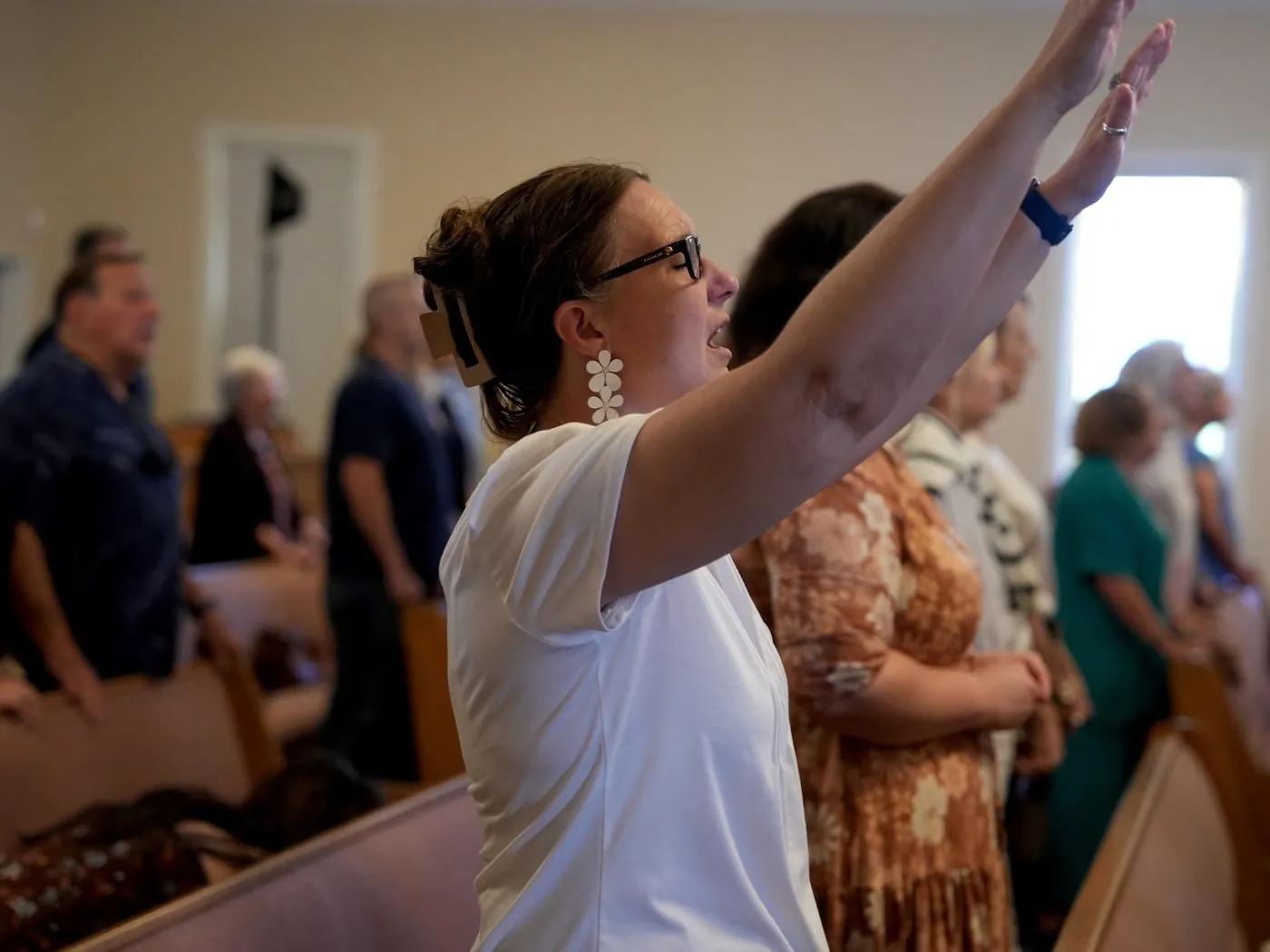 A woman singing with one hand raised during worship, others standing in the pews around her