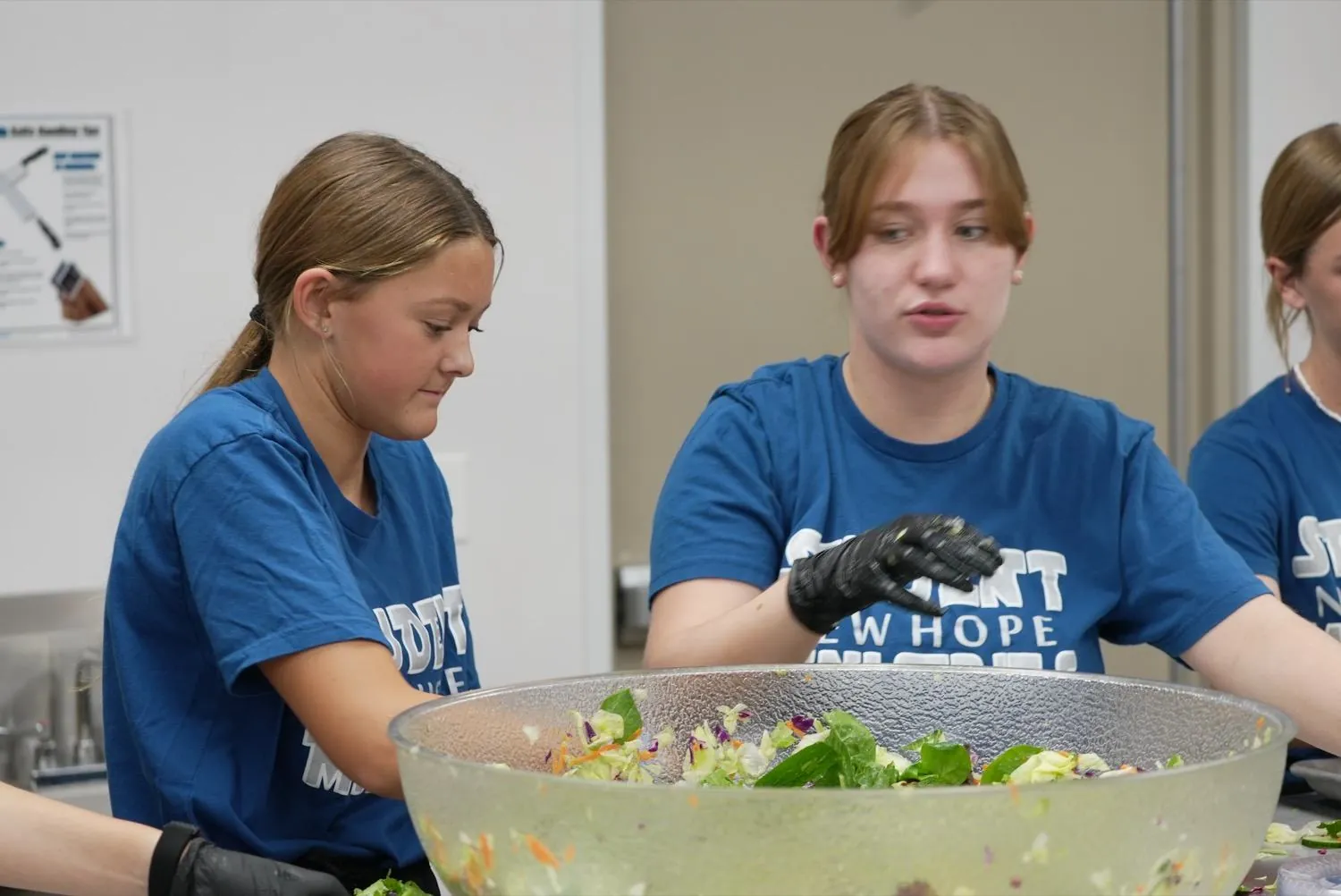 Student volunteers in New Hope ministry shirts tossing a large bowl of salad in the church kitchen