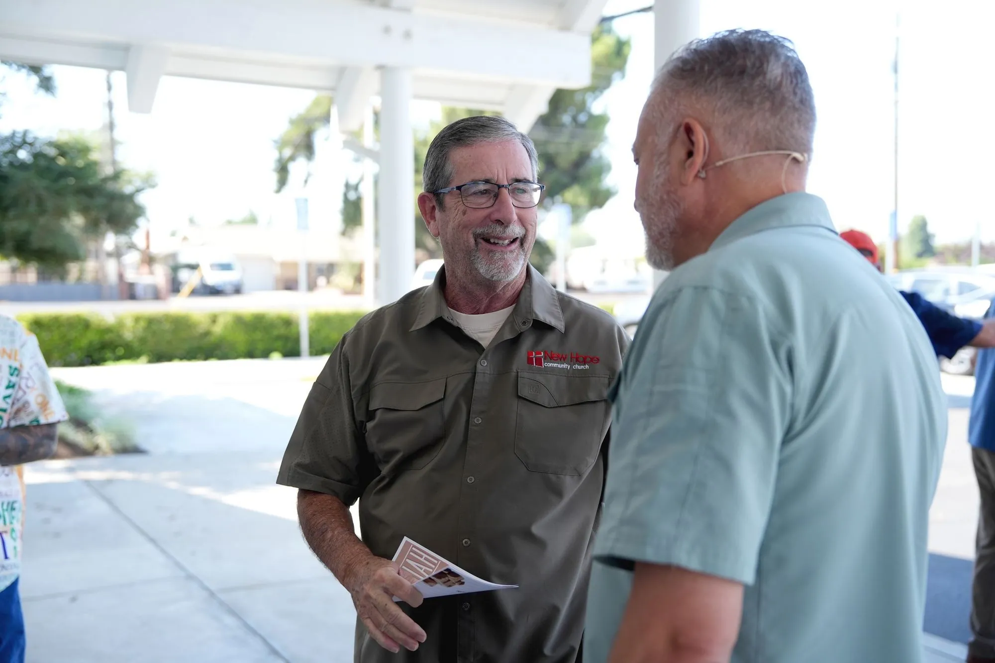 Two men talking under the entry canopy after a Sunday service, one wearing a New Hope shirt and holding a bulletin