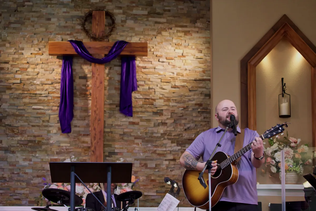 A worship leader singing with an acoustic guitar beside the wooden cross draped in purple on Easter Sunday 2026