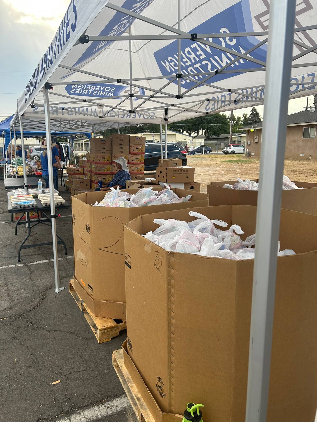 Large cardboard bins filled with bagged food standing under a Sovereign Ministries canopy