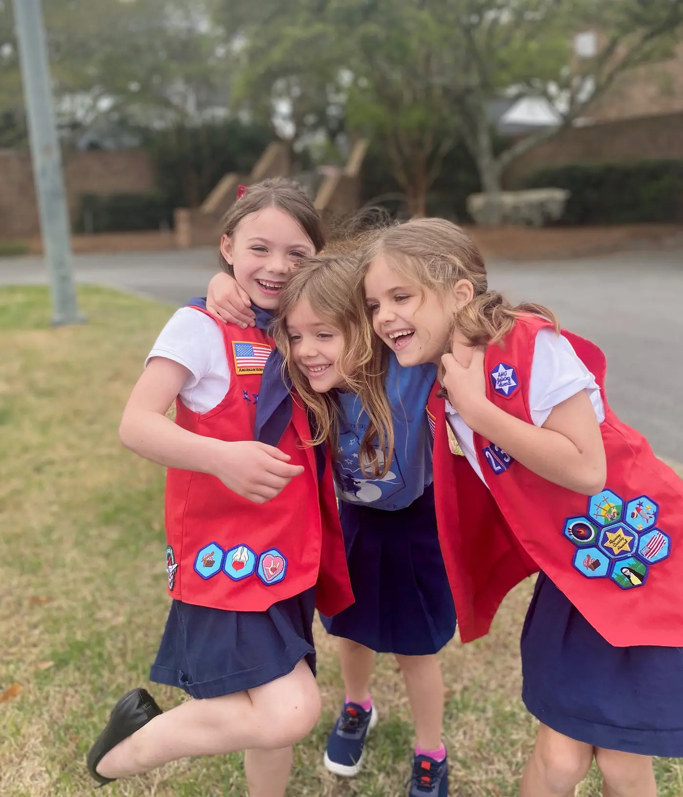 Three American Heritage Girls in badge sashes laughing with their arms around each other