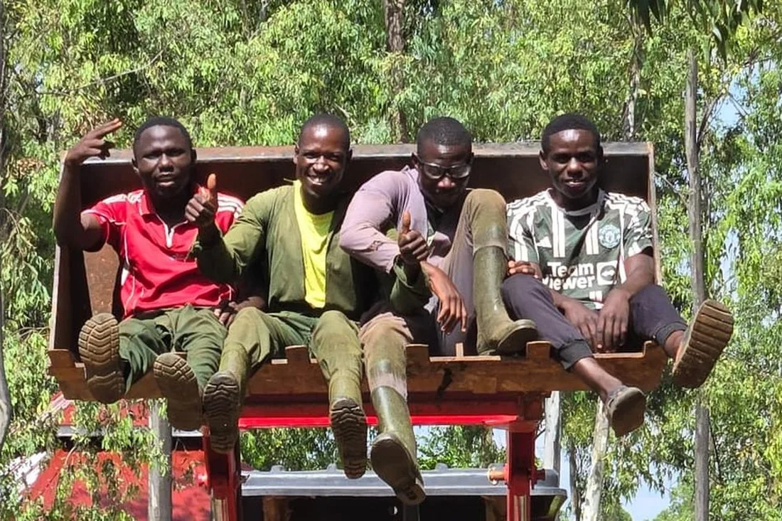 Four farm workers sit together on a red tractor attachment under the trees, smiling.