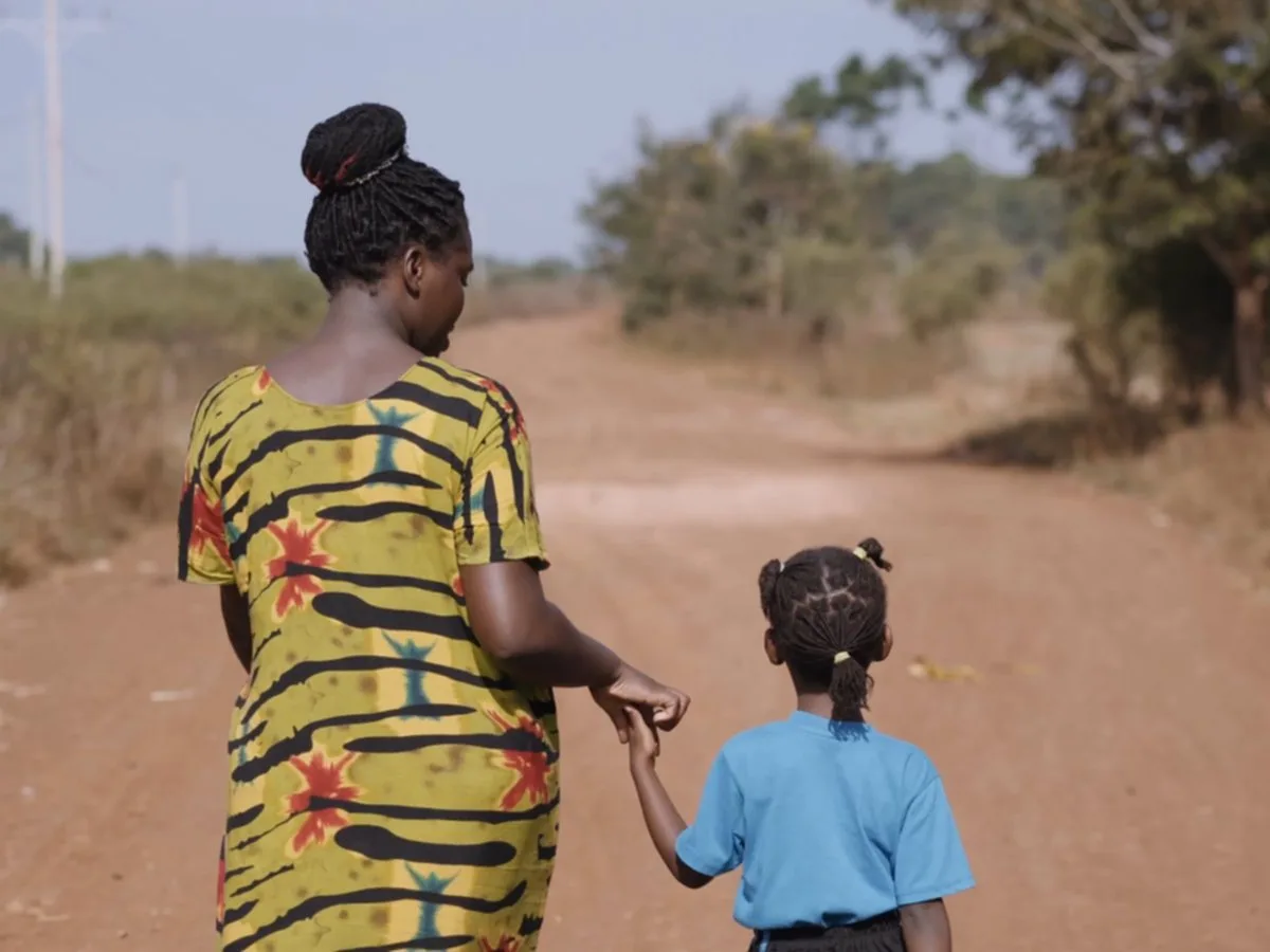 A woman and a young girl walk hand in hand down a red dirt road in Bulesa, seen from behind.