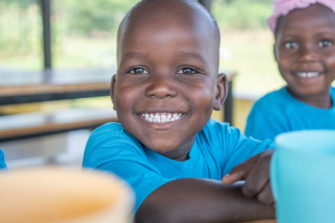 A smiling preschooler in a blue uniform at a classroom table.