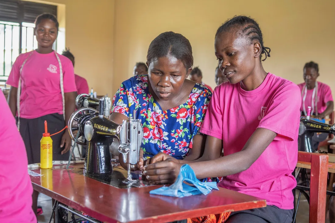 Girls in pink shirts learn to sew at a treadle sewing machine with their instructor.