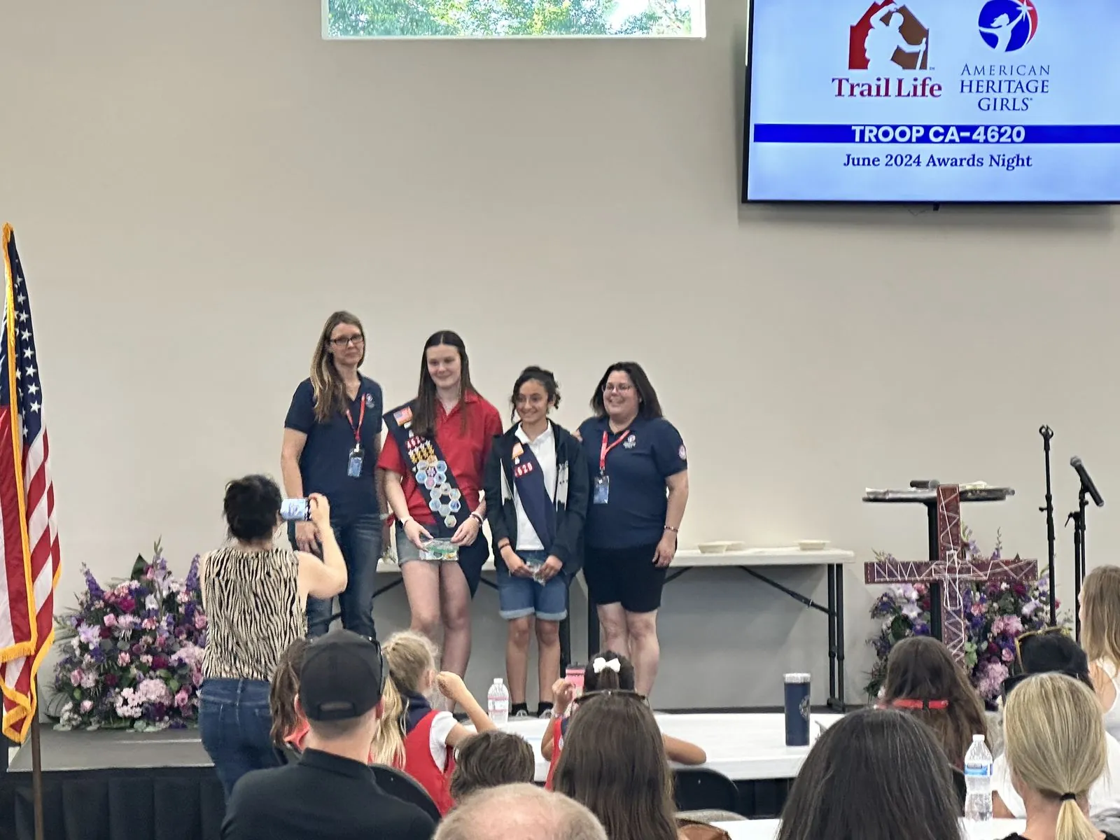 Two American Heritage Girls in their sashes on stage with two adult leaders at the June 2024 awards night for New Hope's Trail Life and American Heritage Girls troops, families watching