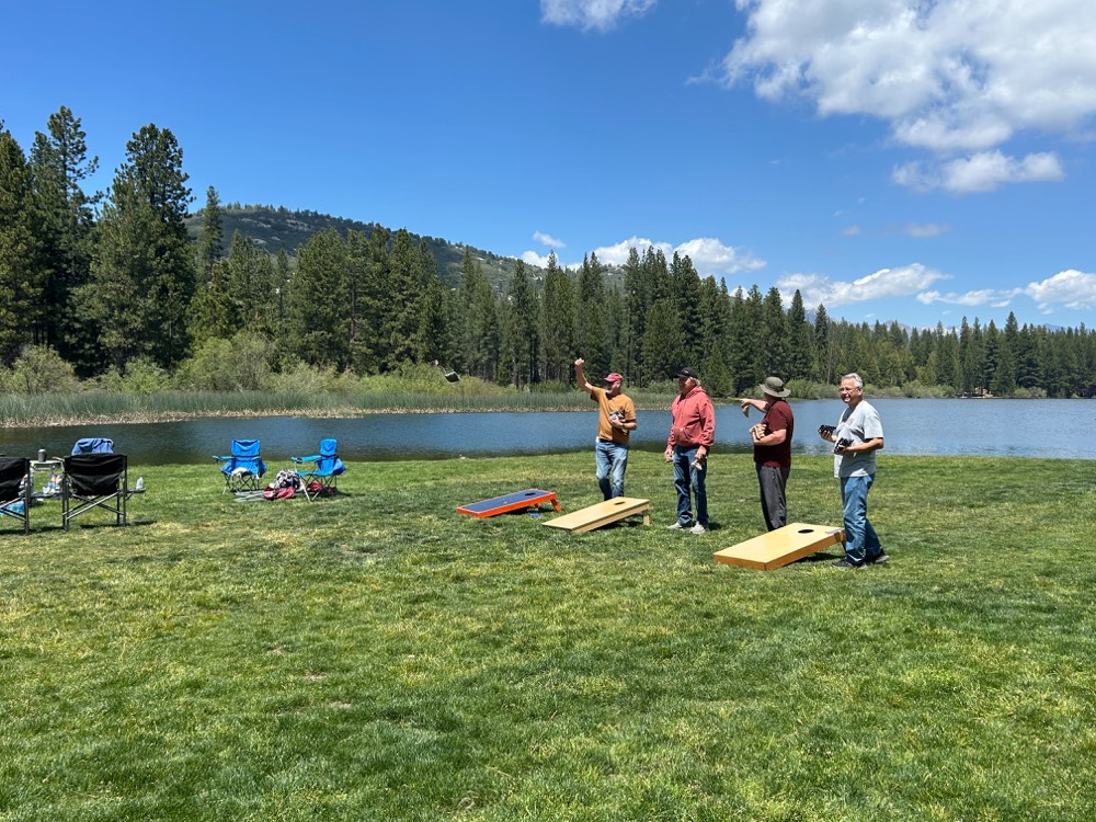 Four husbands playing cornhole at the Hume Lake waterfront on a sunny afternoon, with the lake and Sierra mountains in the background