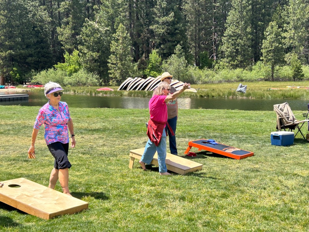 A wife mid-throw at a cornhole game with the lake and canoes behind her
