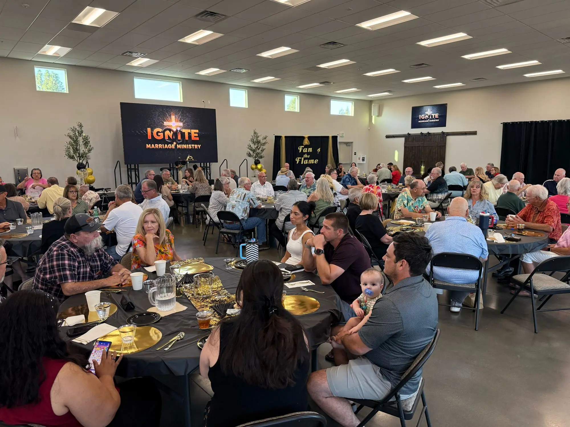 A full room of couples at round tables during the Ignite Marriage Ministry Fan the Flame dinner, with the Ignite screen and stage in the background