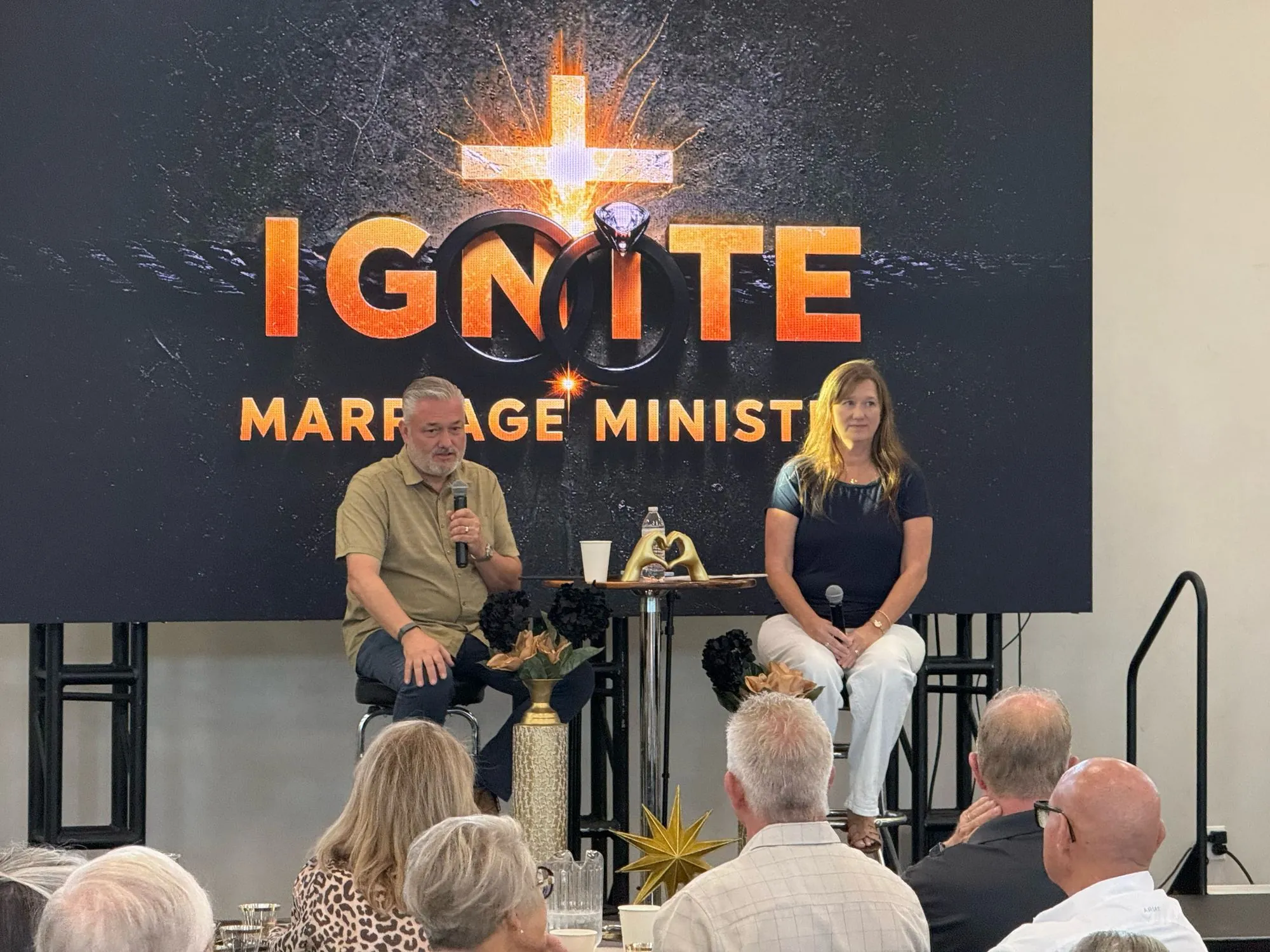 A husband and wife seated on stage sharing with the room in front of the Ignite Marriage Ministry screen