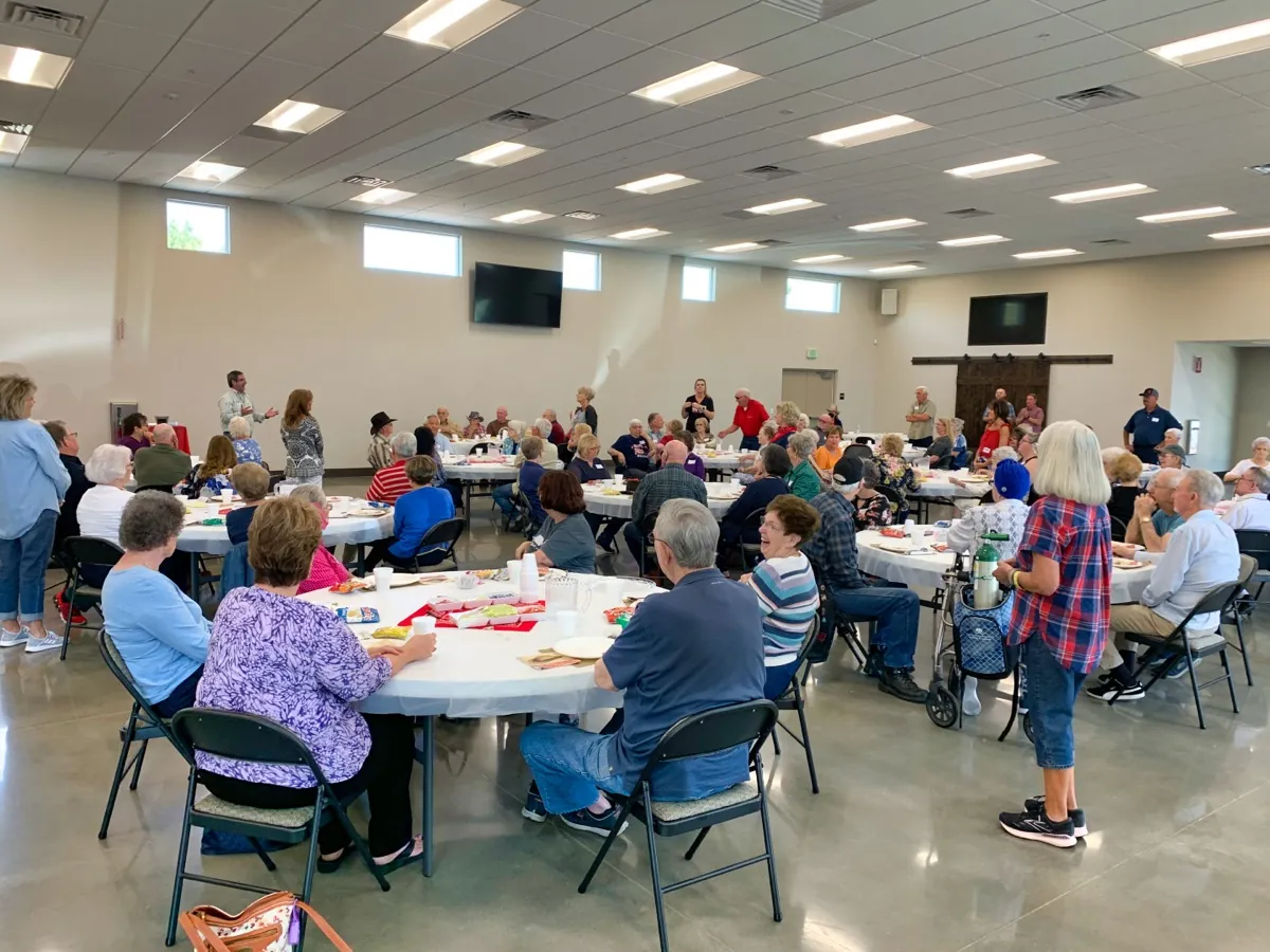 A wide view of New Hope seniors gathered around round tables for the monthly Senior Lunch in The Barn