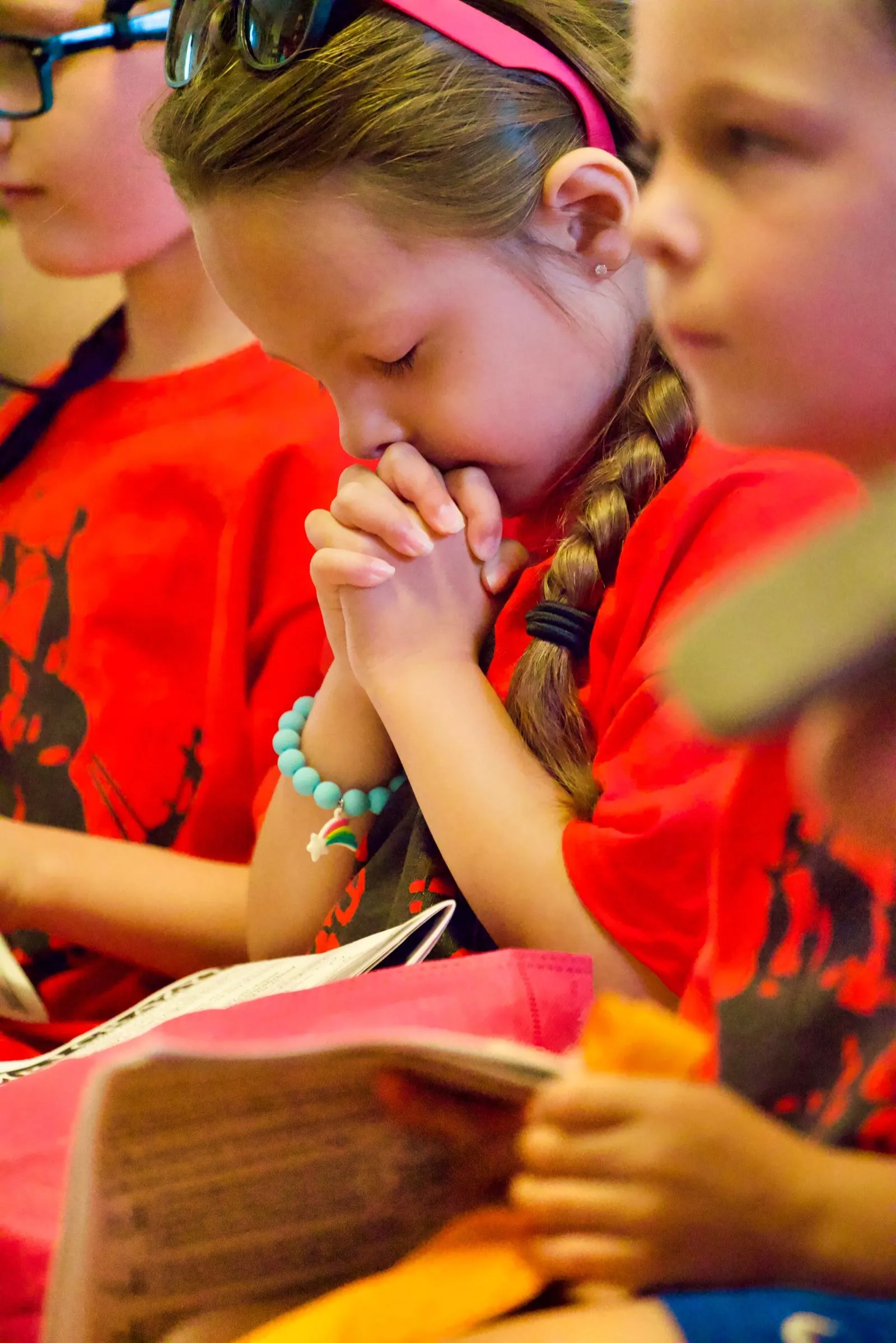 A New Hope girl in a red VBS shirt with hands folded in prayer, a children's Bible open beside her