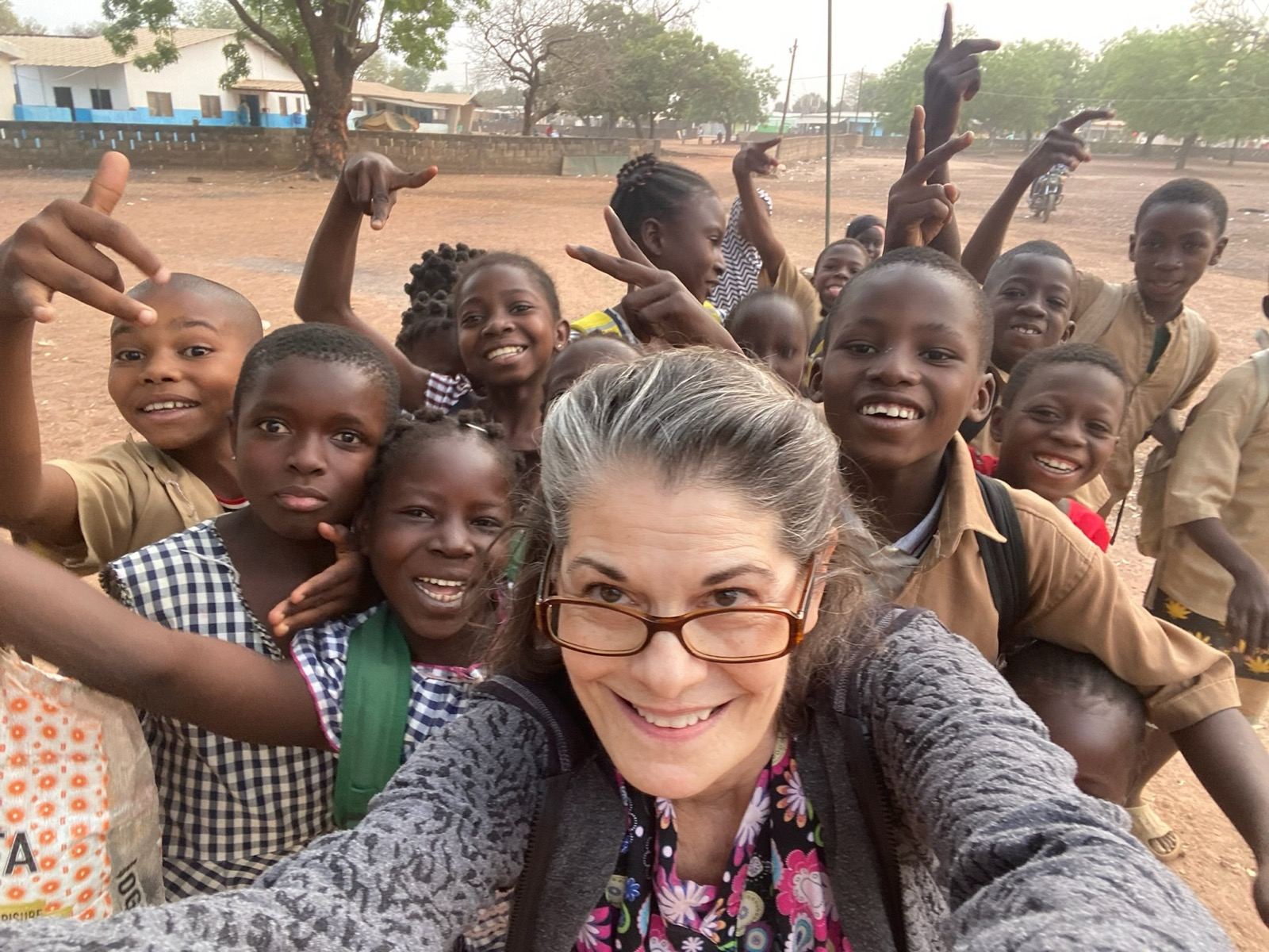 A woman takes a selfie surrounded by smiling schoolchildren in a red-earth schoolyard in Ivory Coast, many of them raising their hands