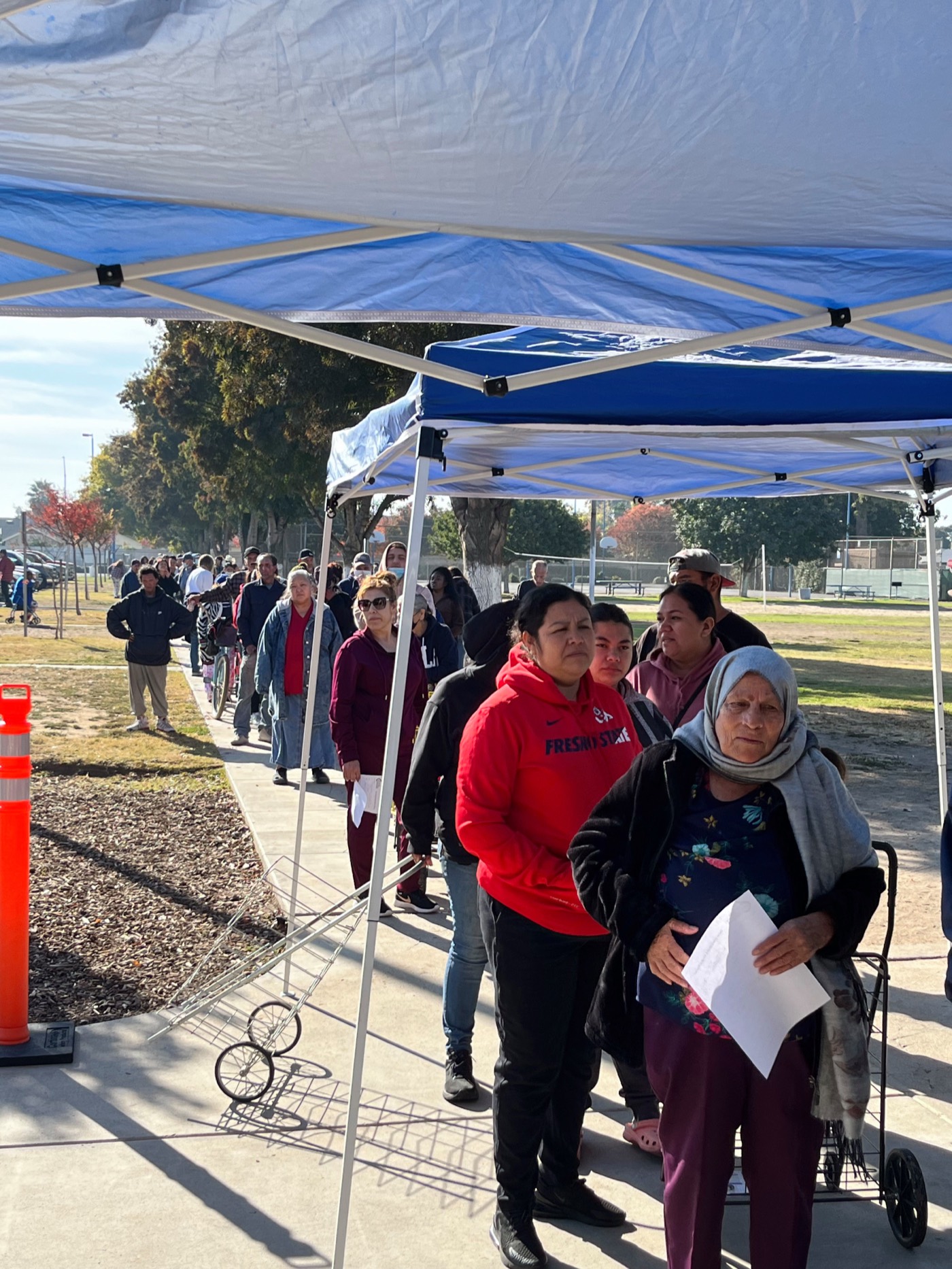 A line of neighbors waiting under the blue Sovereign Ministries canopy at the Calwa food pantry on a sunny weekday morning