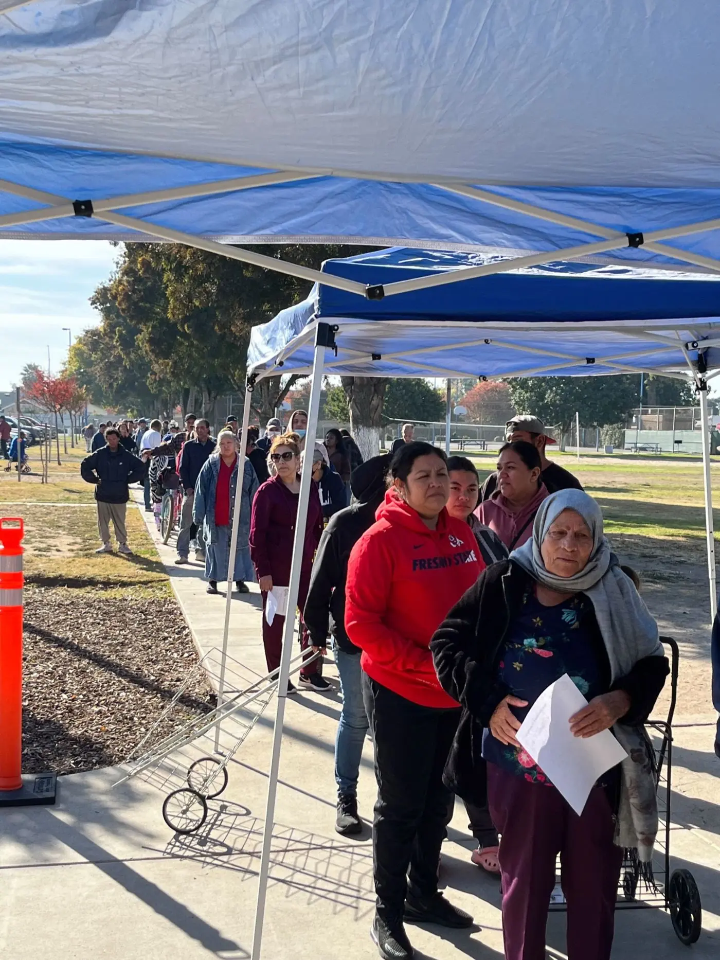 A line of neighbors waiting under the blue Sovereign Ministries canopy at the Calwa food pantry on a sunny weekday morning