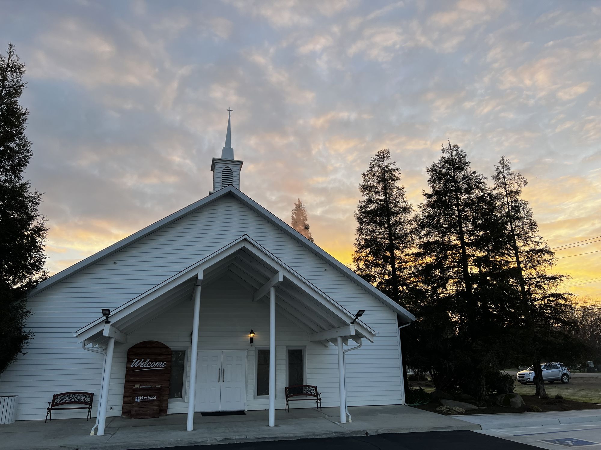 New Hope's Clovis church building at sunrise, gold and pink clouds behind the steeple, with a wooden Welcome sign and two benches beside the front doors