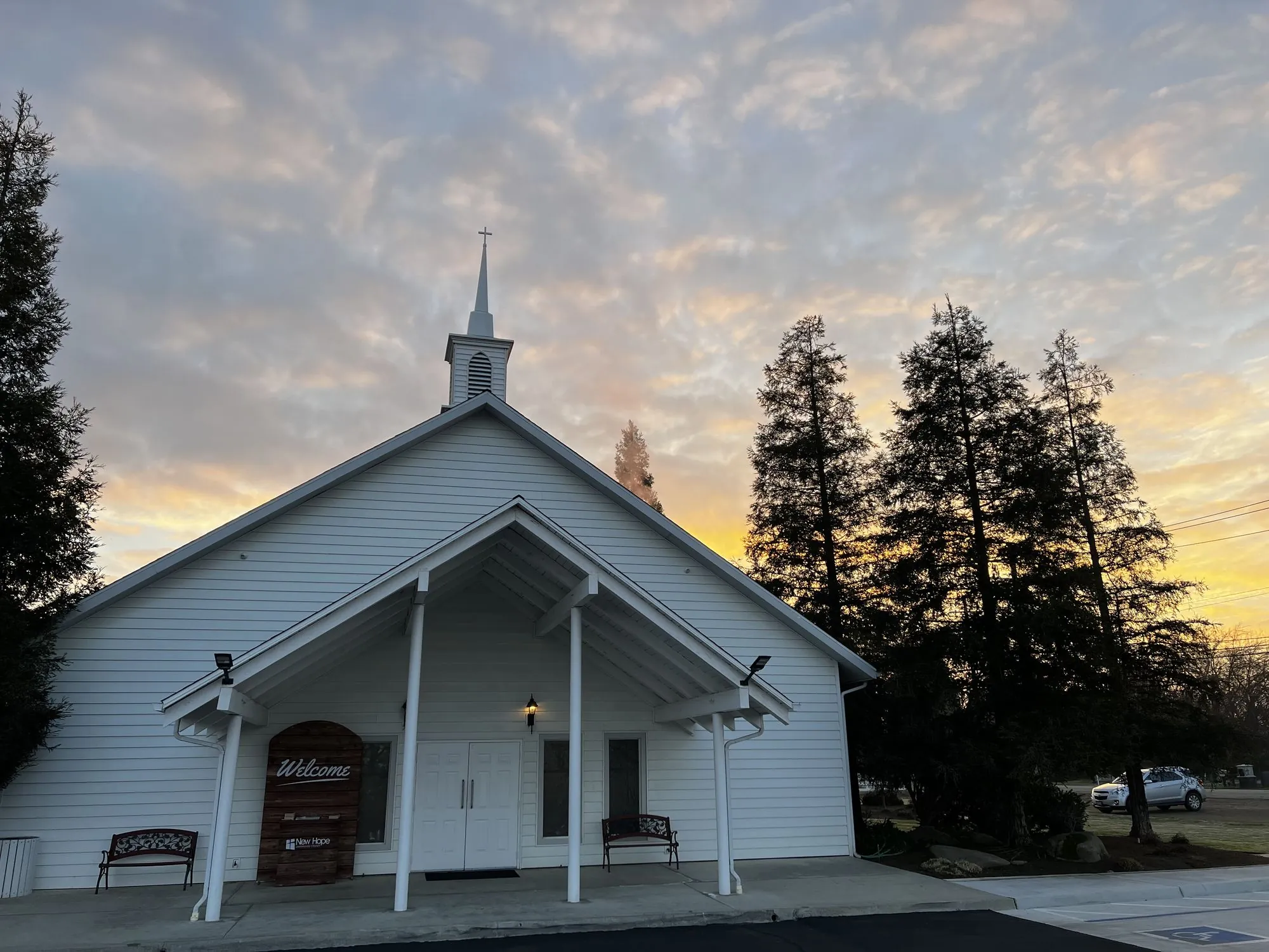 New Hope's Clovis church building at sunrise, gold and pink clouds behind the steeple, with a wooden Welcome sign and two benches beside the front doors