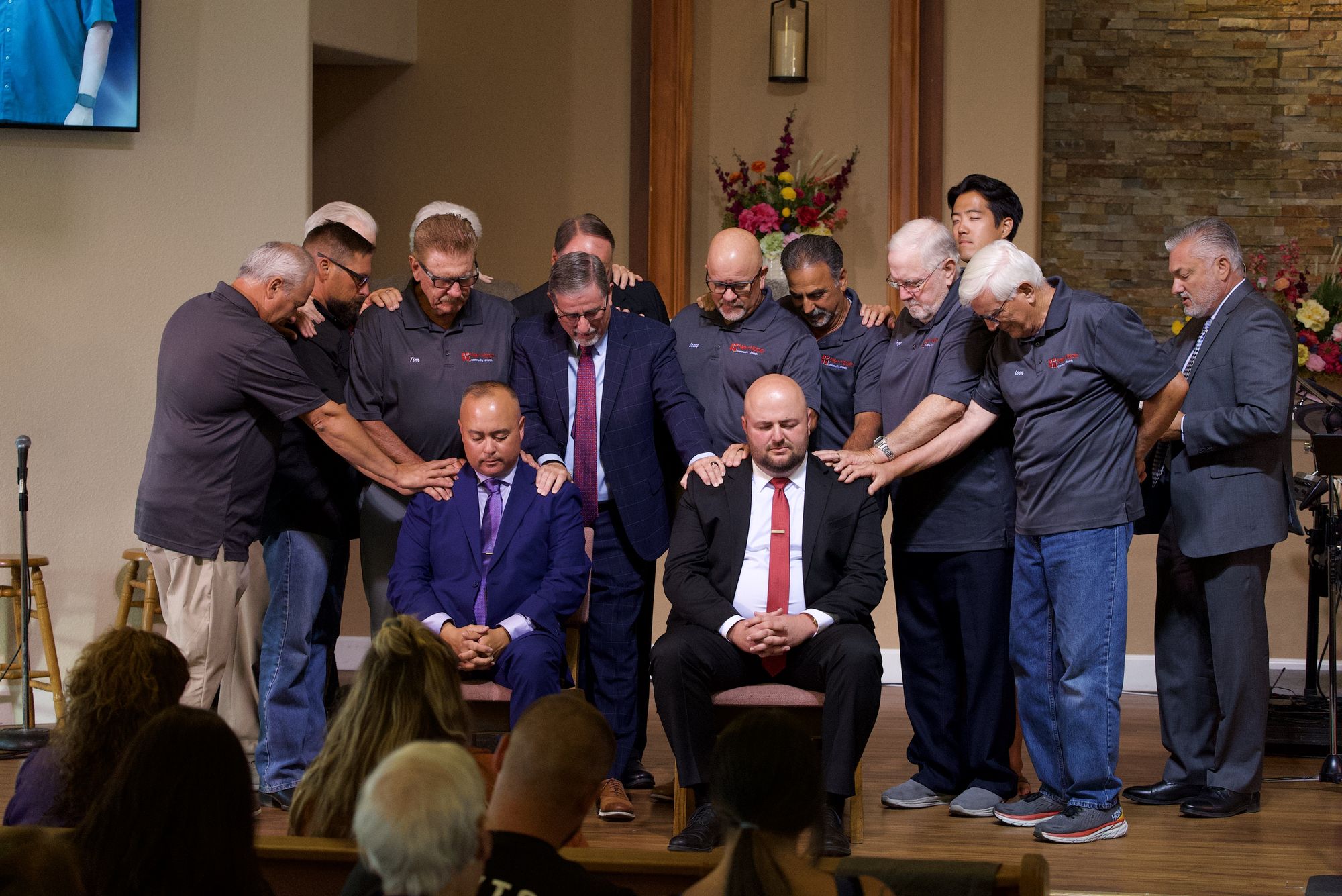 New Hope elders and pastors gathered on the sanctuary platform, hands laid on the shoulders of two seated men with every head bowed, during an ordination prayer