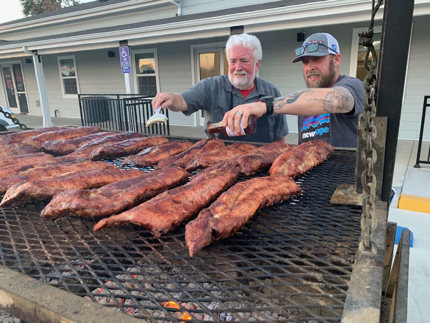Two men brushing sauce on racks of ribs over an open grill at a New Hope men's barbecue