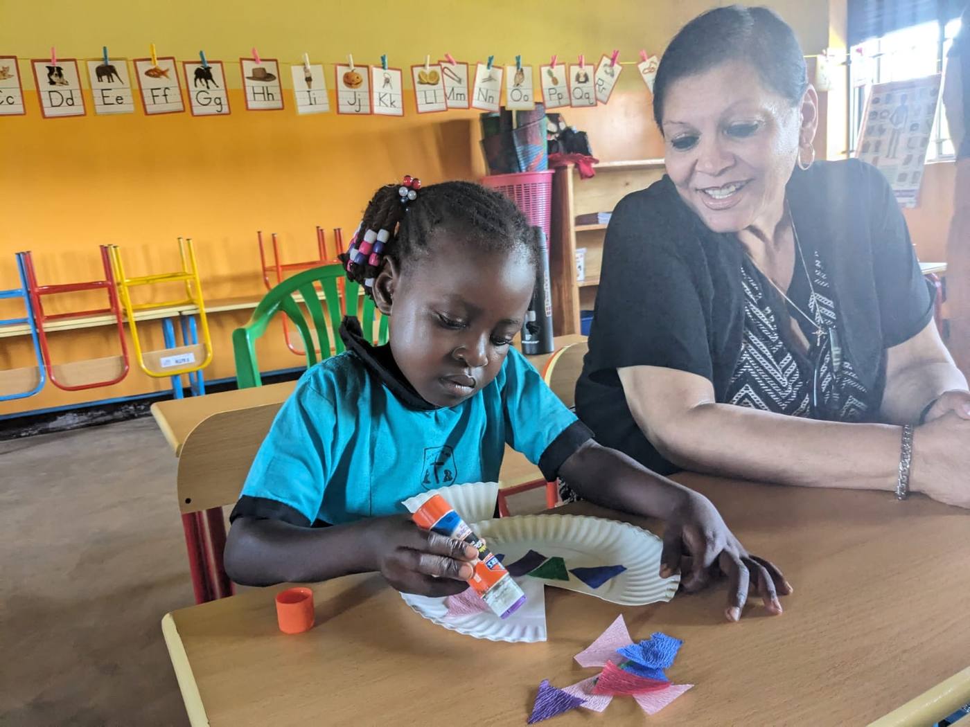A New Hope team member helping a young girl with a craft in a brightly painted classroom in Uganda