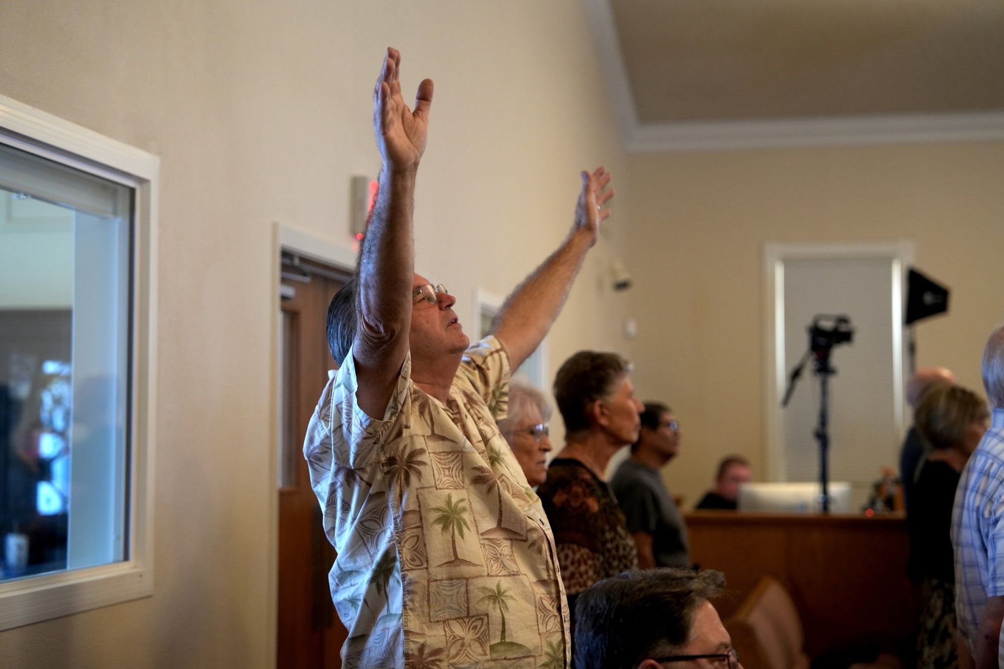 A man with both arms raised in worship during a service at New Hope in Clovis, with the video team recording from the booth behind him