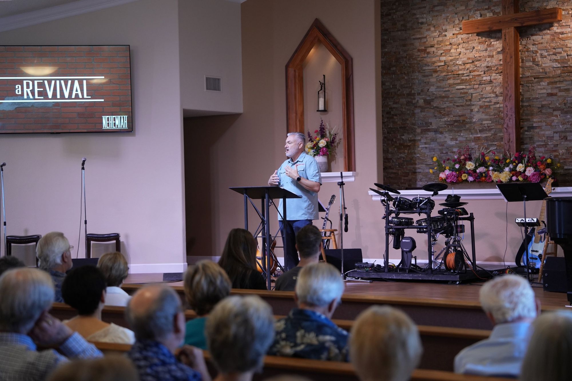 Pastor preaching with the sermon title graphic on screen and the stage audio and drum equipment the media team runs each Sunday