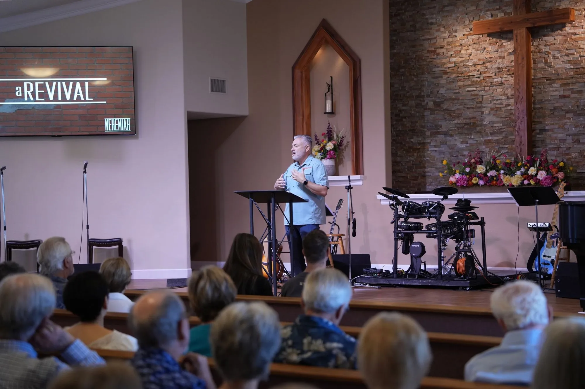 Pastor preaching with the sermon title graphic on screen and the stage audio and drum equipment the media team runs each Sunday