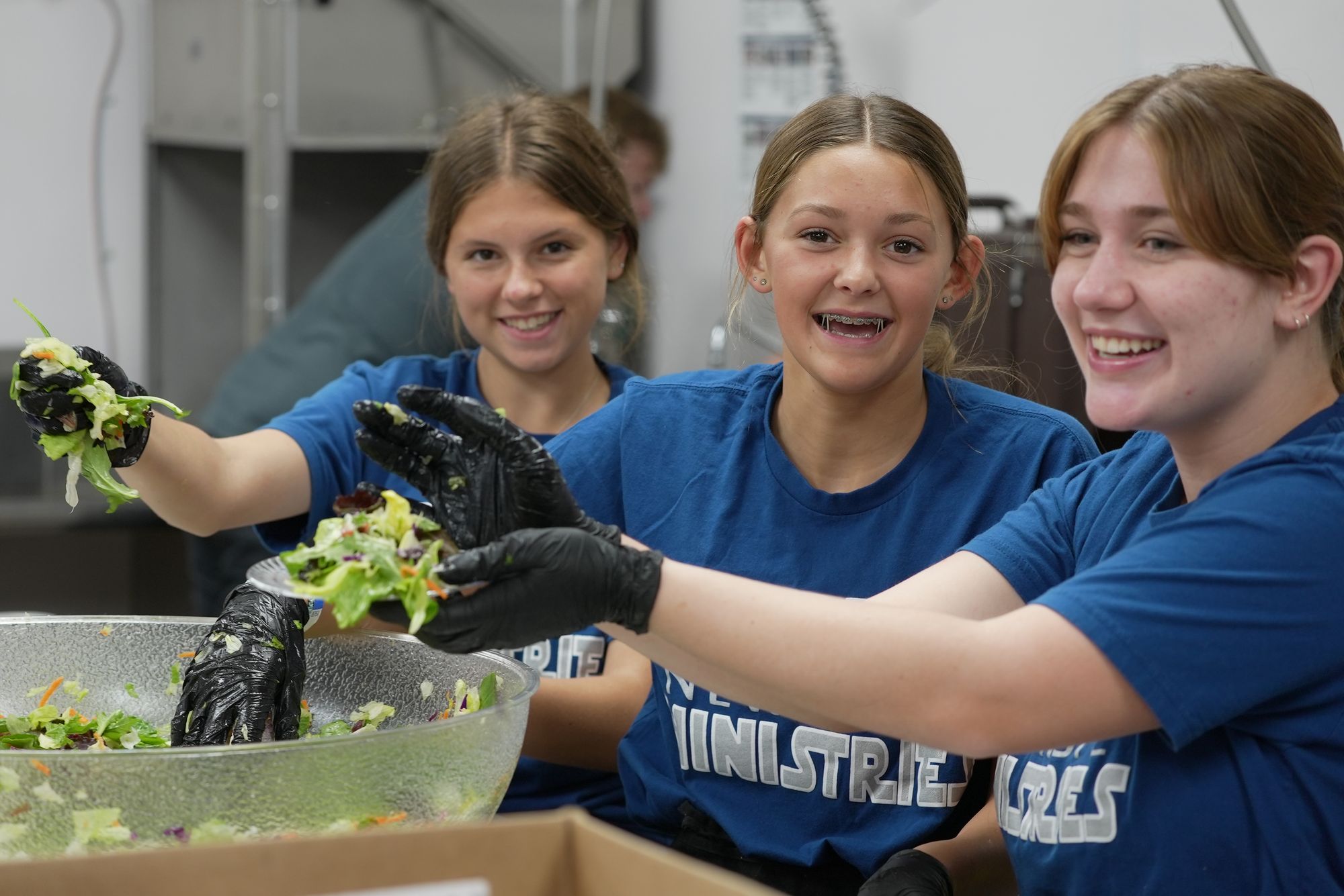 Three teenage volunteers in blue New Hope t-shirts and food-service gloves tossing a large bowl of salad in the kitchen, smiling at the camera