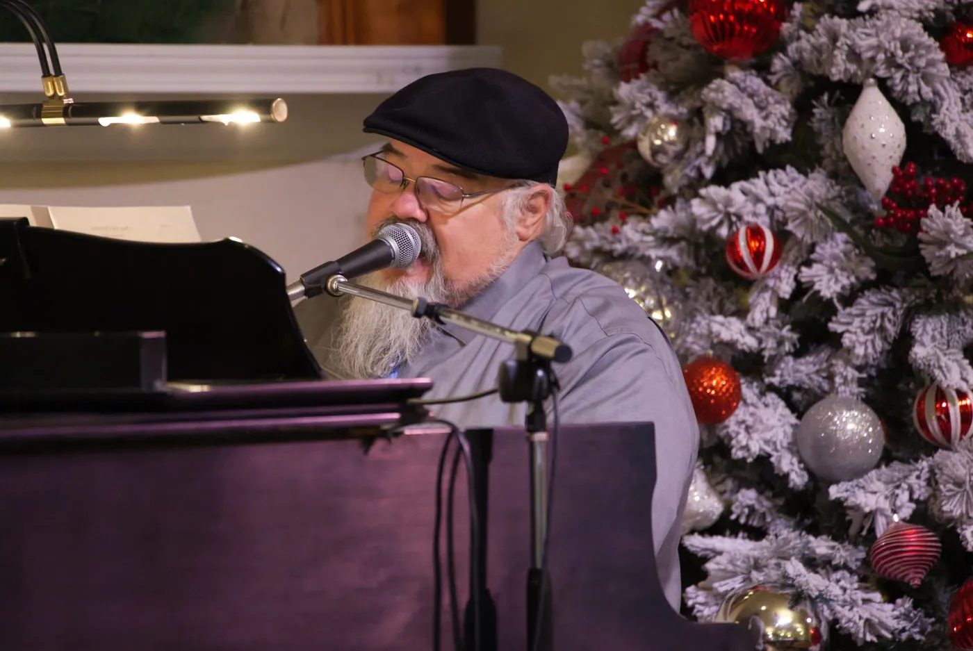 An older man in a flat cap singing into a microphone at the piano, beside a flocked tree hung with red and silver ornaments