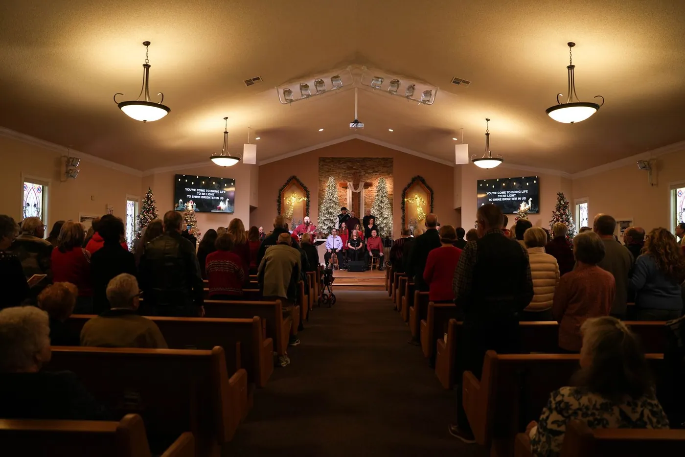 The New Hope sanctuary from the back of the center aisle during a Christmas service, people standing in the pews and Christmas trees at the front