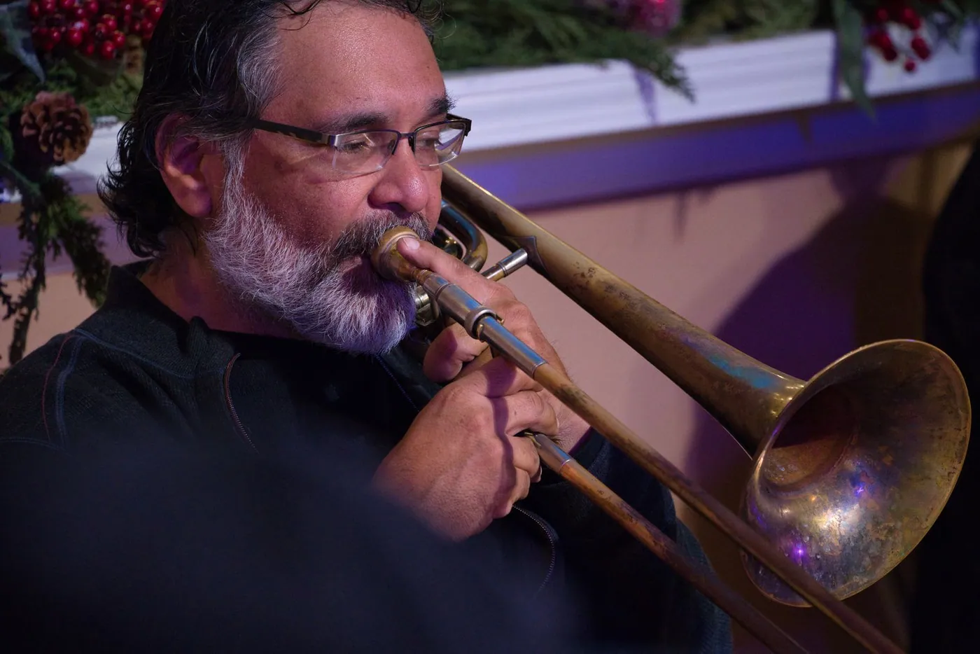 A bearded man in glasses playing trombone, pine garland and red berries on the ledge above him
