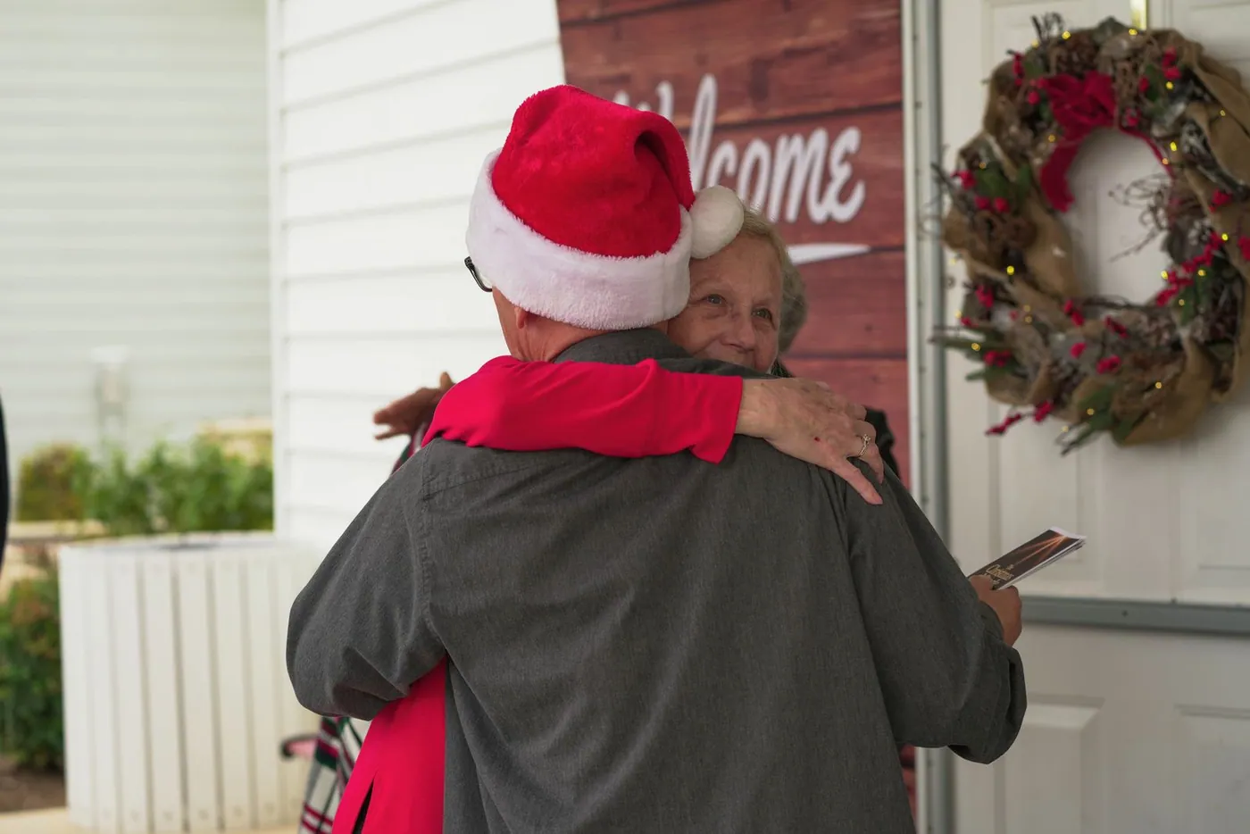 A man in a Santa hat hugging a smiling silver-haired woman at a New Hope door hung with a Christmas wreath, beside a Welcome sign