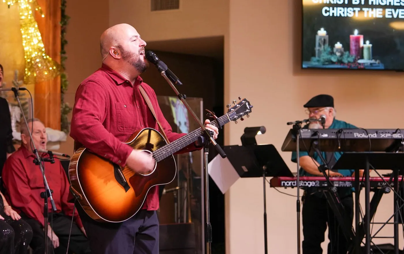 A man in a red shirt singing and playing acoustic guitar at a New Hope Christmas service, a keyboard player behind him
