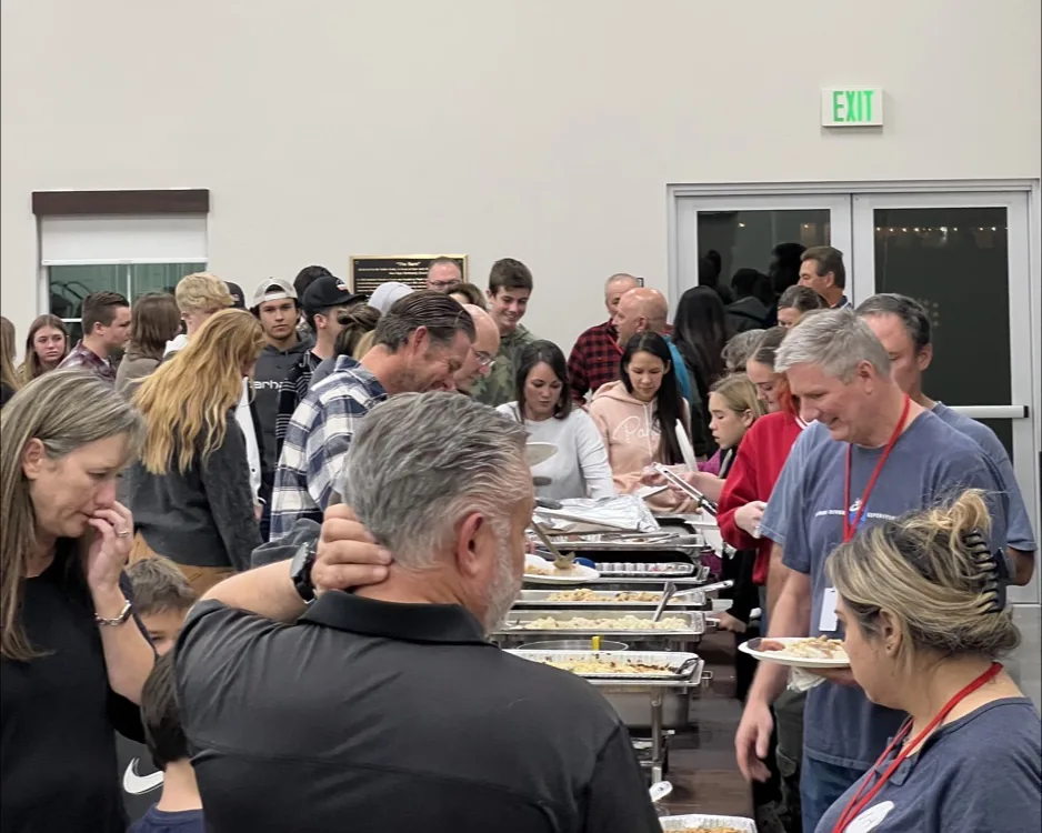 A mixed-age crowd lined up at a long buffet table of chafing dishes in the New Hope fellowship hall, filling their plates