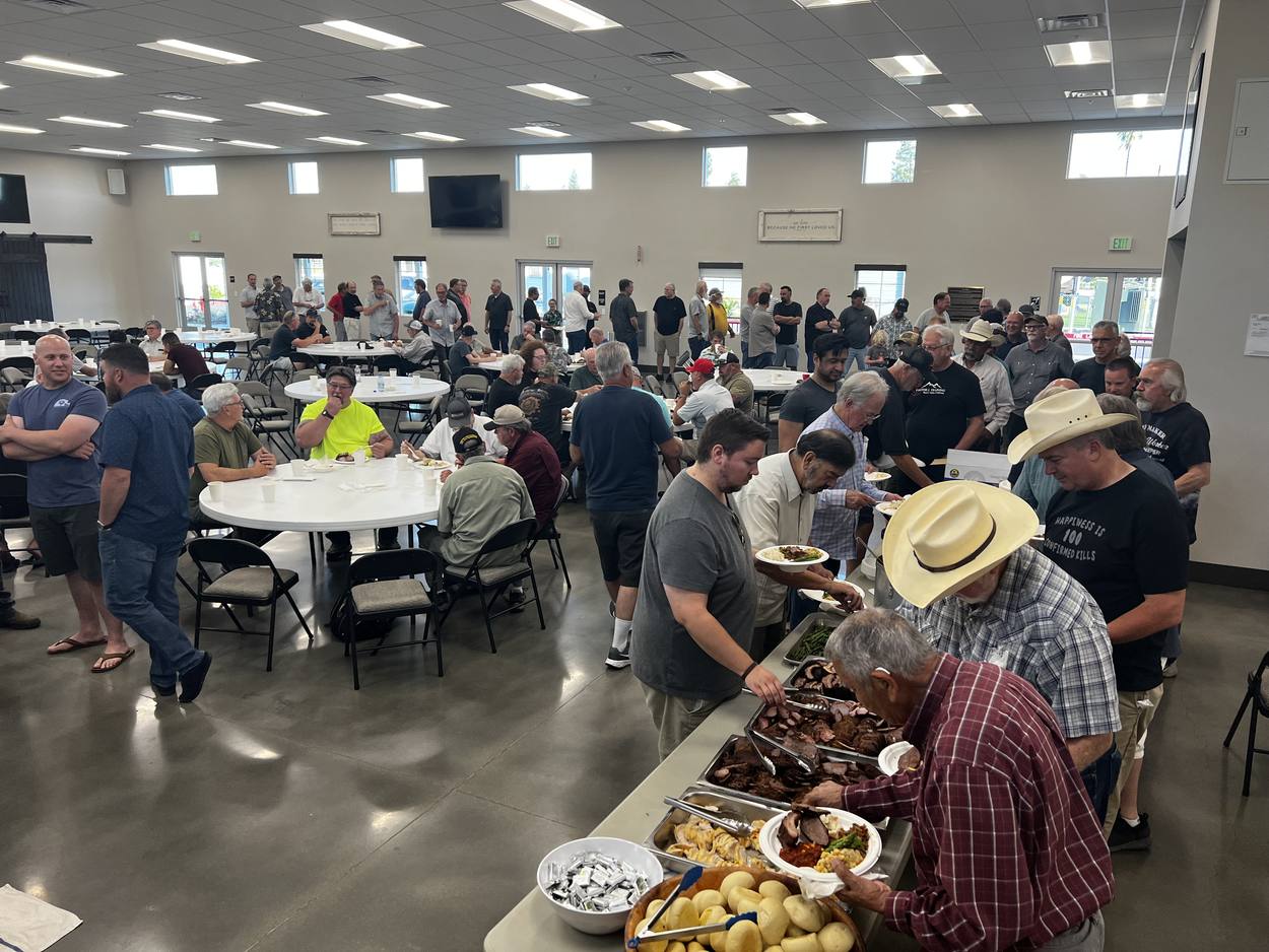Men of varied ages gathered around a long serving table loaded with platters of grilled meat and potatoes in the New Hope fellowship hall