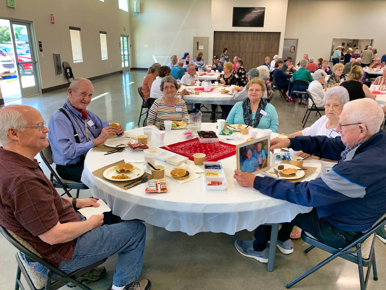 Adults seated around a round table at New Hope in conversation over plates of food, red napkins on the table and the busy hall behind them