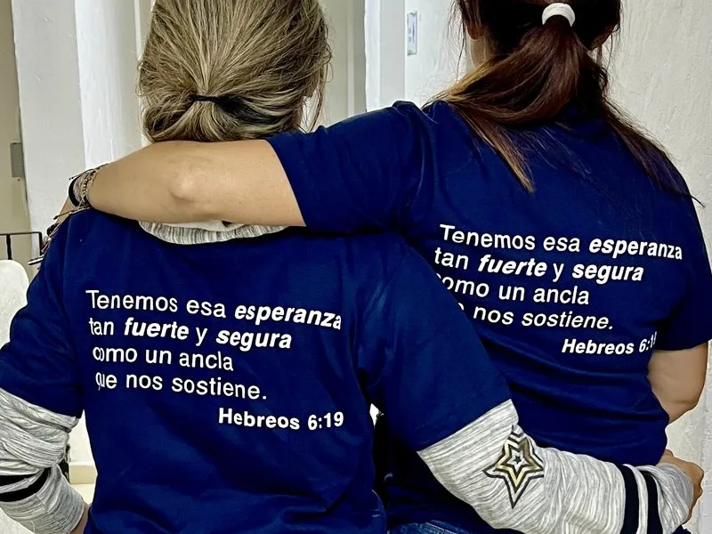 Two women walk arm in arm, seen from behind, in matching navy shirts printed with Hebrews 6:19 in Spanish.