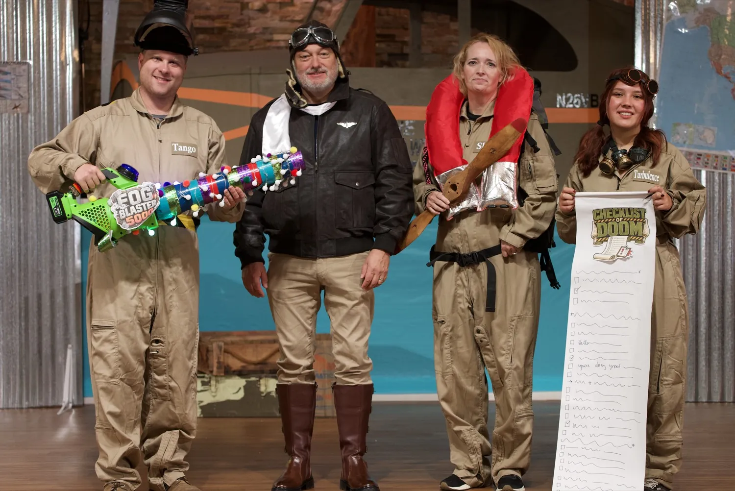 The Skybound cast in costume on the set: four crew members in flight suits, one with the light-up Fog Blaster and one holding the captain's checklist, beside the captain in his aviator jacket