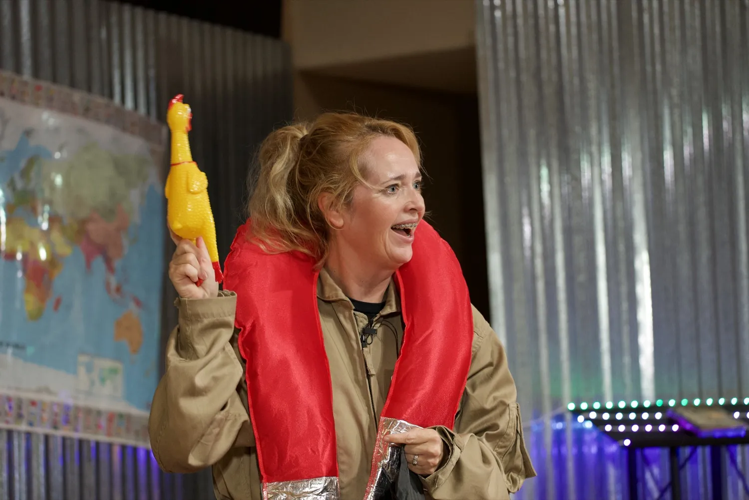 Skye in a flight suit and red life vest, holding up a rubber-chicken prop mid-scene