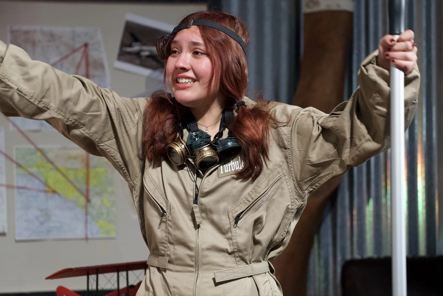 Turbulence in a flight suit and aviator goggles, arms thrown wide mid-scene