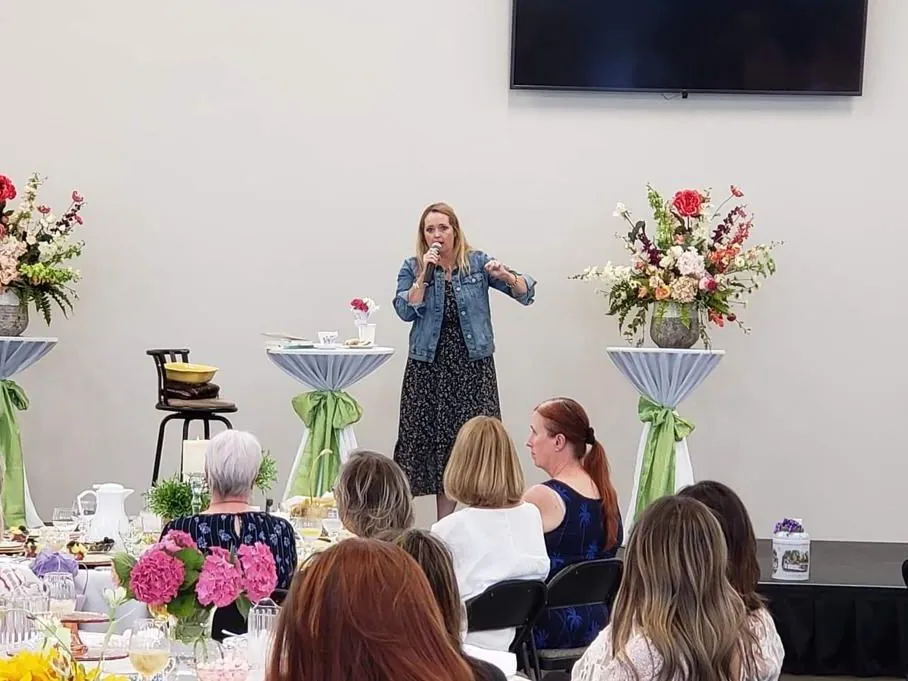 A speaker with a microphone addressing women seated at decorated tables at a New Hope women's gathering